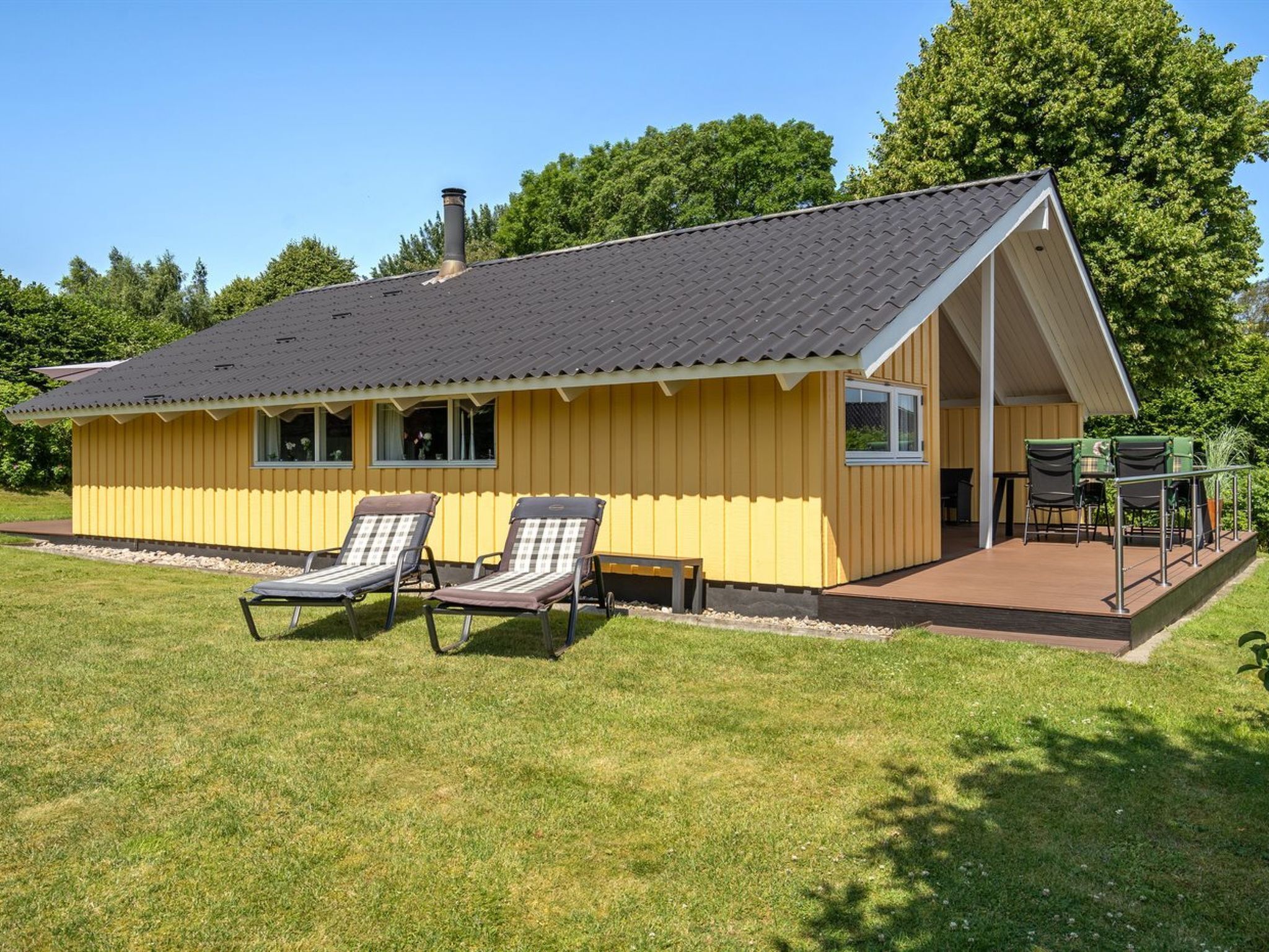 Ein gelbes Gebäude mit dunklem Dach, zwei Liegestühlen und einer Terrasse mit Stühlen und einem Tisch.