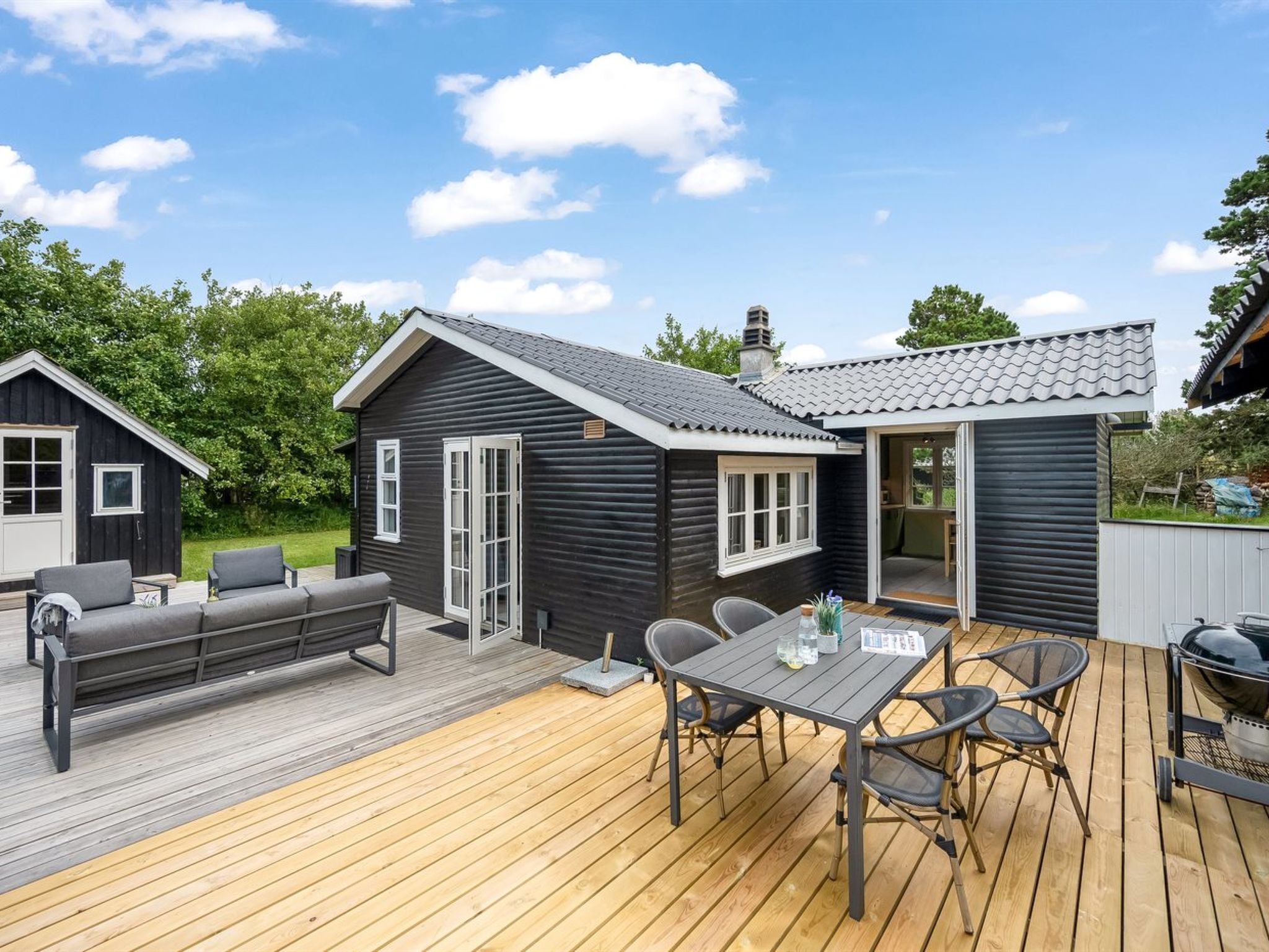 Eine Holzterrasse mit Esstisch und Stühlen, einem Grill und einer kleinen Sitzecke mit Sofa und Sesseln.