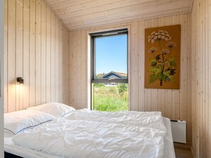 Et soveværelse med trævægge, en seng med hvide sengetøj, et vindue der viser et hus, og et plantemotiv på en vægmonteret billedramme.