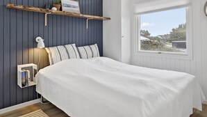 Un dormitorio con cama, lámpara de noche, estante con una foto y una planta, y ventana con vistas al exterior.