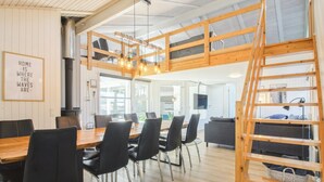 Amplio espacio tipo loft con mesa de comedor de madera, sillas negras, cama en alto con escalera de madera y un gran ventanal que proporciona luz natural.