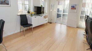 Een moderne woonkamer met een flatscreen televisie, een bureau en een stoel. Er zijn ramen met zonwering, een deur naar buiten en een lijst met een foto aan de muur.