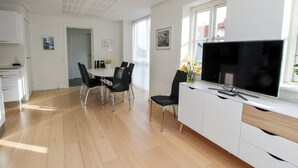 Een moderne woonkamer met een flatscreen televisie, een eethoek met een witte tafel en zwarte stoelen, en een keuken met witte kasten en een houten vloer.