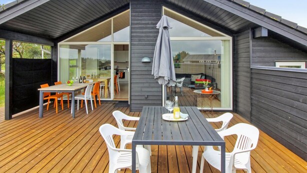A modern house with a wooden deck, a dining area with a table and chairs, and a large glass door leading to a kitchen. - "Alma" - 500m from the sea by Interhome (Løkken)