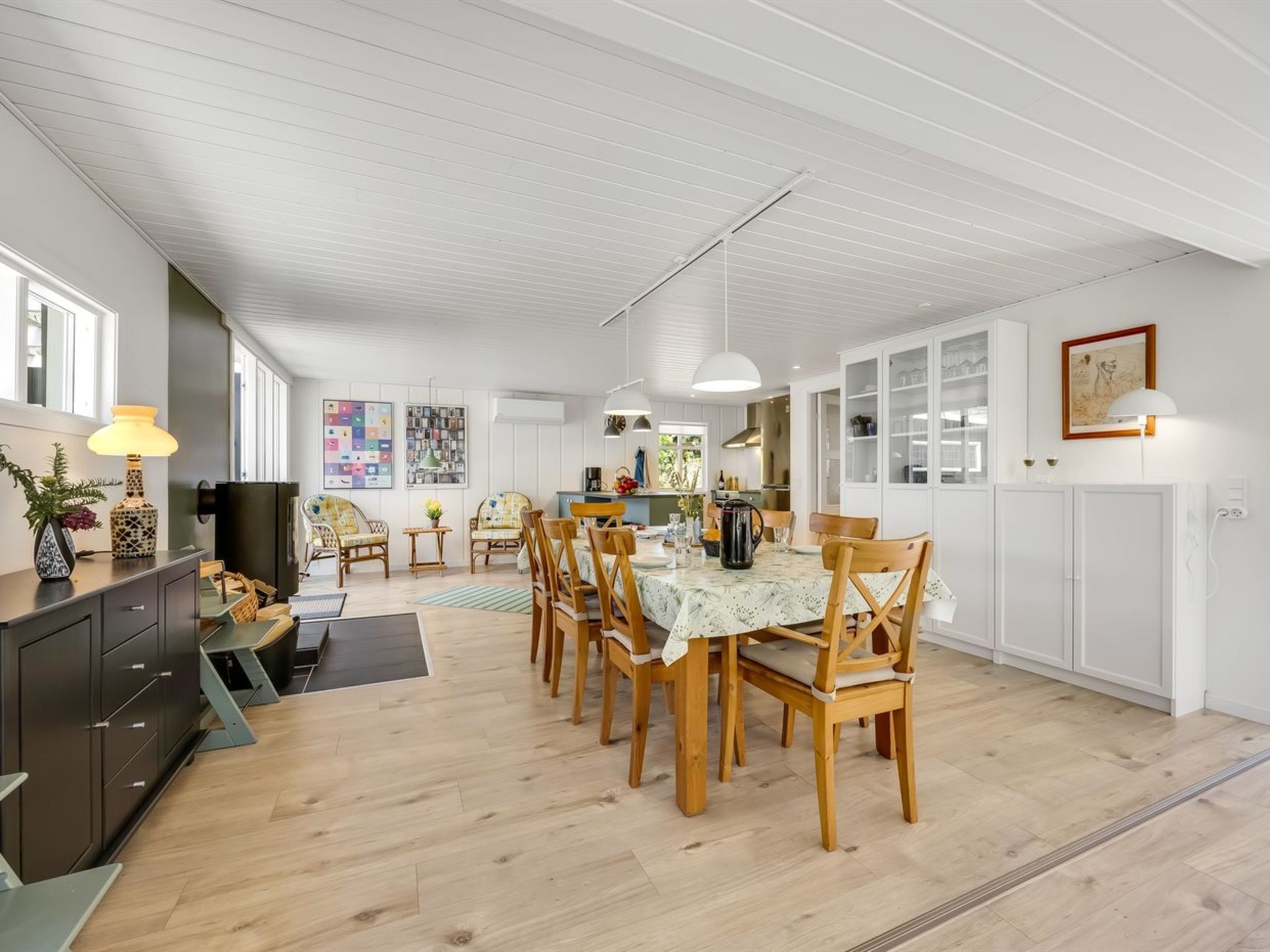 A spacious dining area with wooden chairs and a table, white walls, and a kitchen in the background.