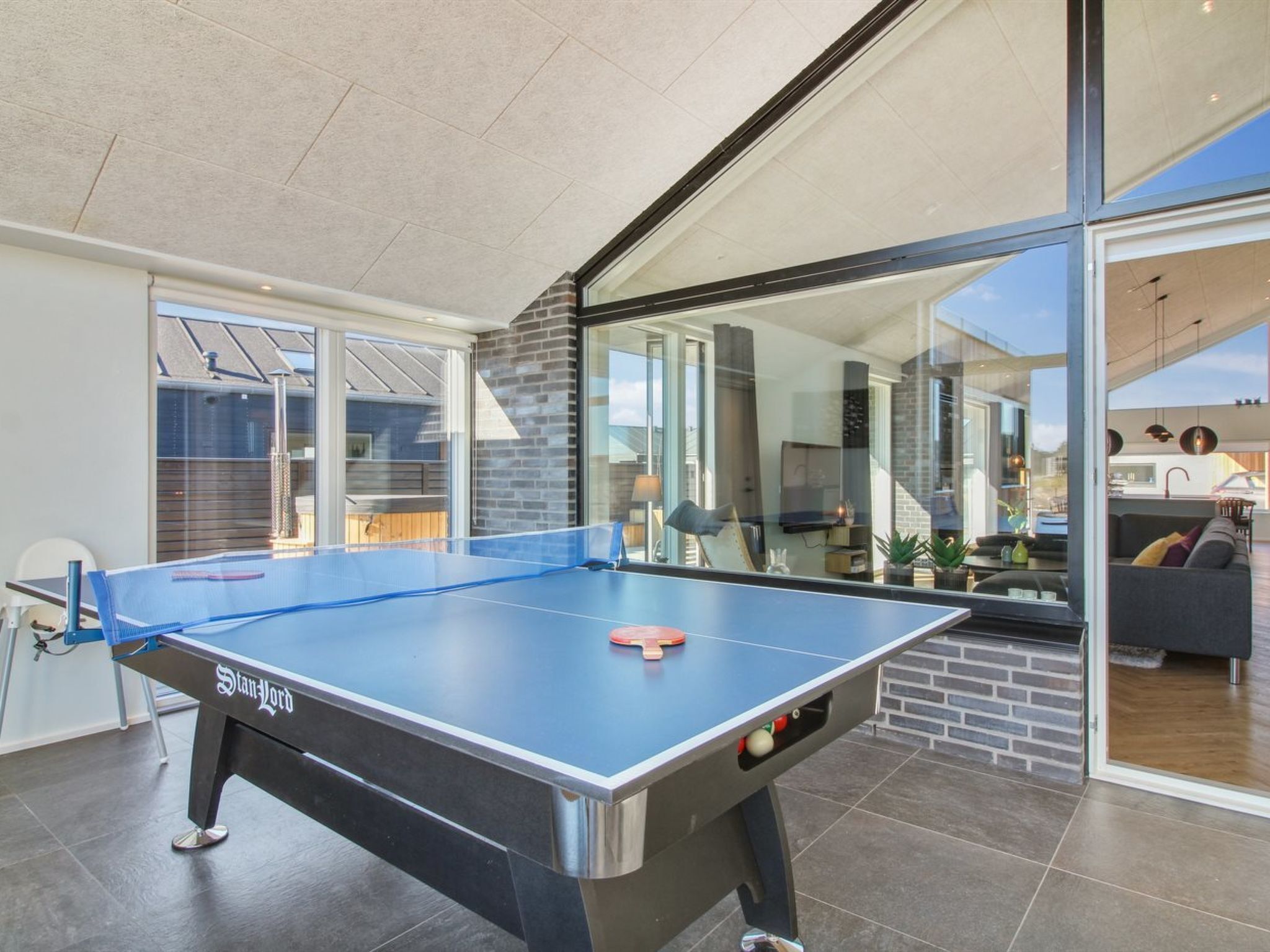A ping pong table in a modern indoor space with large windows and a view of a living area.