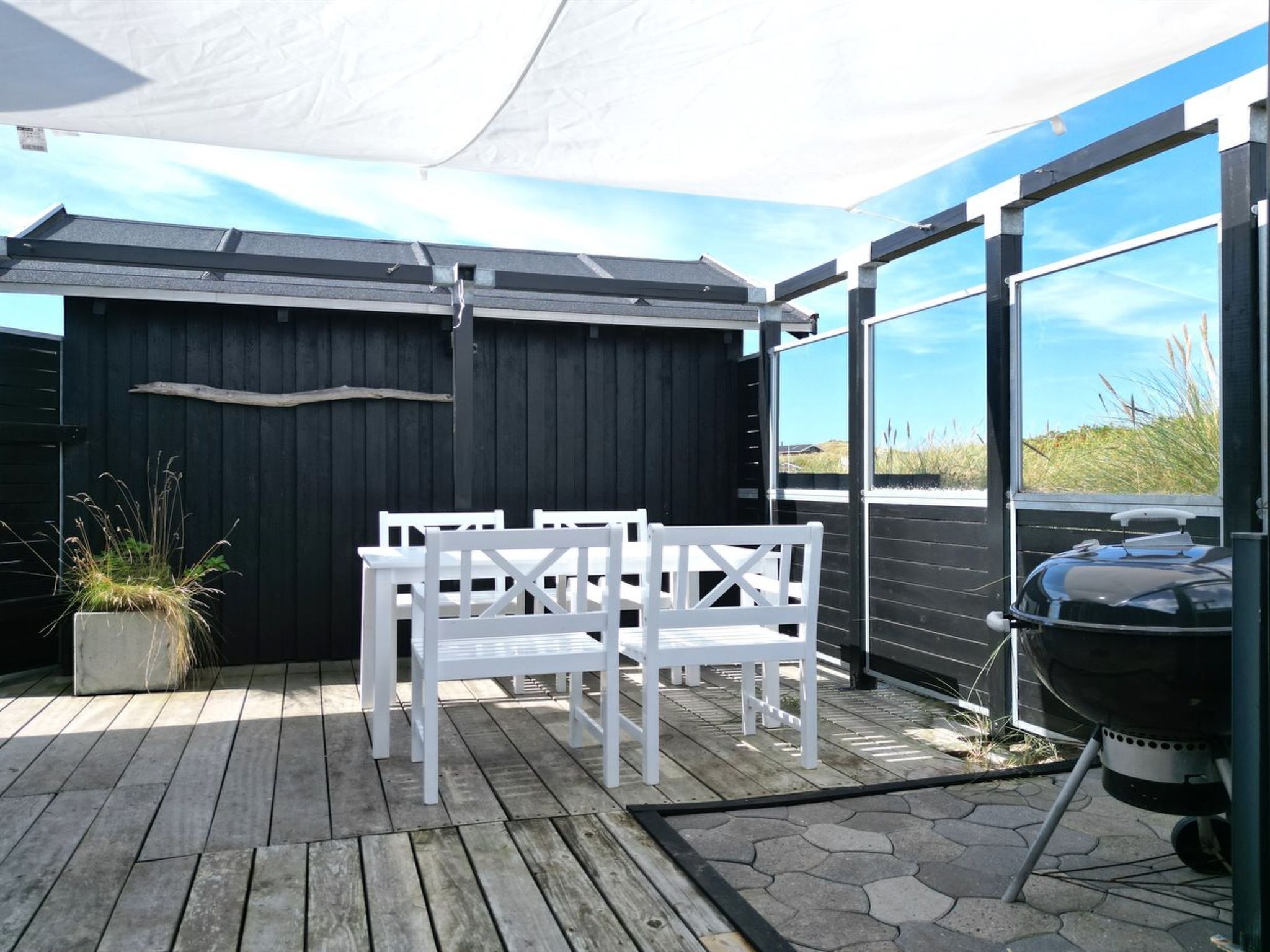 Ein überdachter Patio-Bereich mit weißer Gartenmöbeln, einem Grill und Blick auf den Strand durch große Fenster.