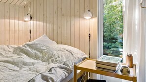 Een slaapkamer met een plafond van houten latten, een bed met witte beddengoed, twee wandlampen en een raam met uitzicht op groen.