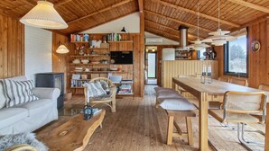 A cozy cabin interior with a wooden ceiling, a living area with a sofa and coffee table, a dining area with a wooden table and chairs, and a bookshelf filled with books and decorative items. - "Nestori" - 950m from the sea by Interhome (Løkken)