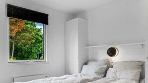 Een eenpersoonsbed met hoofdeinde, een nachtkastje met lamp, een raam met zonwering en uitzicht op bomen.