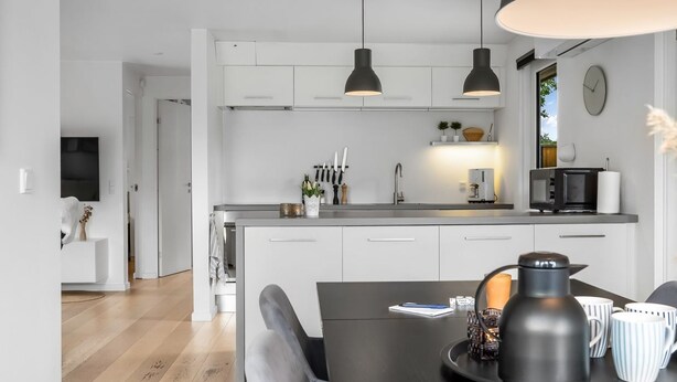 Een moderne keuken met witte kasten, een centraal eiland, hanglampen, een eethoek met een zwarte tafel en een zichtbare klok aan de muur.