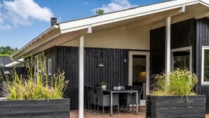 Een houten terras met een tafel, stoelen, een deur naar binnen en twee plantenbakken met planten.