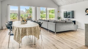 Ein geräumiges Wohnzimmer mit hoher Decke, einem großen Sofa, einem Esstisch mit Stühlen und einem an der Wand befestigten Fernseher.