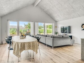 A spacious living area with a high ceiling, a large sofa, a dining table with chairs, and a TV mounted on the wall.