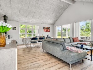 A spacious living area with a high ceiling, large windows, a grey sectional sofa, and a dining area with chairs and a table.
