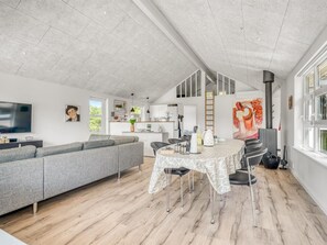 A modern living space with a grey sectional sofa, a dining table with chairs, a kitchen area, and a large window.