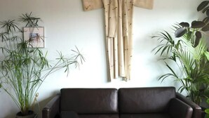 Um quarto com um sofá marrom escuro, uma cortina bege com pequenos desenhos e duas plantas em vasos com folhas longas e finas.