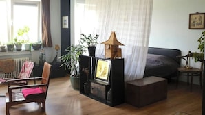 Quarto com cama, poltrona, mesa pequena e diversas plantas em vasos.