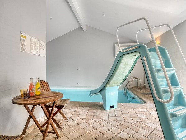 Una piscina con tobogán, una mesa de madera con bebidas y piso de baldosas.