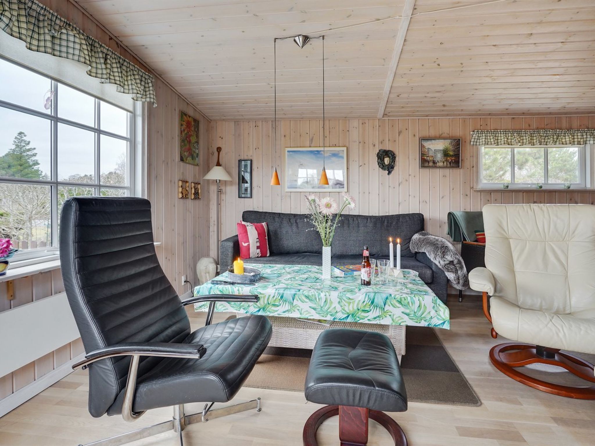 Une pièce chaleureuse avec un canapé, une table à manger recouverte d’une nappe à motifs floraux, un fauteuil en cuir noir, un fauteuil blanc et un plafond en bois.
