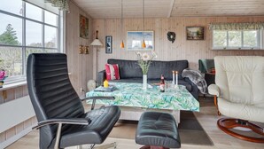 Une pièce chaleureuse avec un canapé, une table à manger recouverte d’une nappe à motifs floraux, un fauteuil en cuir noir, un fauteuil blanc et un plafond en bois.
