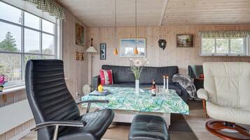 Une pièce chaleureuse avec un canapé, une table à manger recouverte d’une nappe à motifs floraux, un fauteuil en cuir noir, un fauteuil blanc et un plafond en bois.