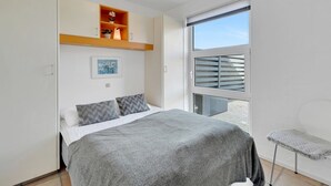 Une chambre à coucher avec un lit, une petite étagère ornée d’objets décoratifs, une fenêtre avec des stores et une chaise.