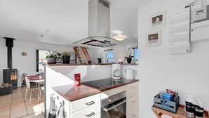 Une cuisine moderne équipée d’un four encastré, d’une cuisinière et d’une machine à café. On y trouve également un coin repas avec une table et des chaises, ainsi qu’un espace de vie agrémenté d’une cheminée.
