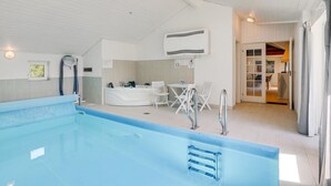 Una piscina interior moderna con agua cristalina azul, un techo iluminado por el sol y un comedor con mobiliario blanco.