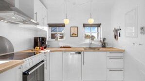 Cocina moderna con armarios blancos, campana extractora de acero inoxidable, horno integrado y encimera de madera. Dos lámparas colgantes sobre el fregadero.