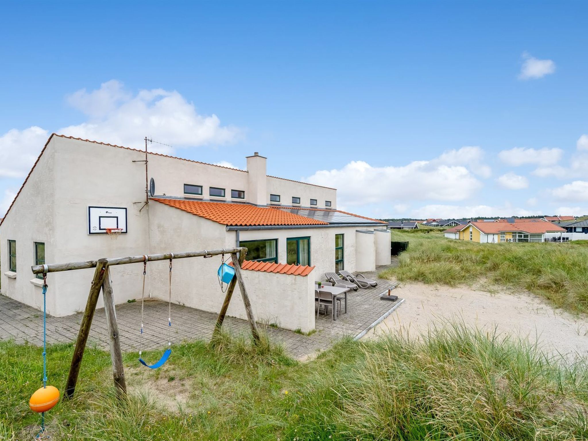Una casa moderna con aro de baloncesto, mobiliario exterior y una zona de arena.