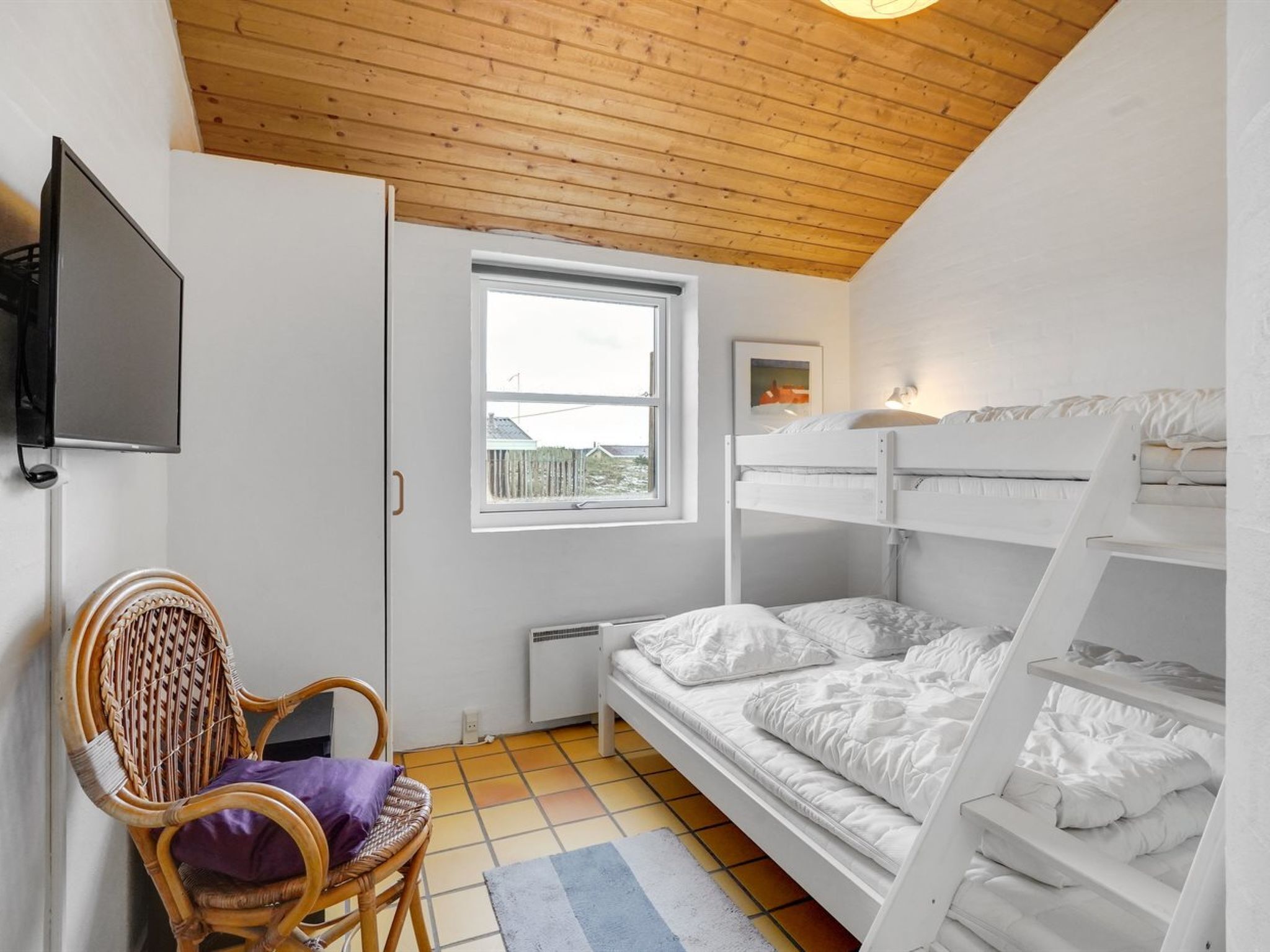 Una stanza con un letto a castello, una televisione fissata al muro, una poltrona in vimini con un cuscino e una finestra vista recinzione.