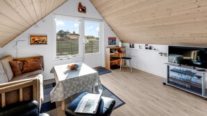 Et hyggeligt loftsværelse med skråvægge, et spisebord med et geometrisk dug, en sofa, et fjernsyn på et stativ og et vindue med udsigt over naturen.
