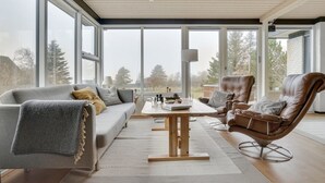 A spacious living room with a grey sofa, a brown leather chair, and a wooden coffee table. Large windows offer a view of trees and a building outside.