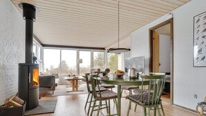 A modern living room with a wood-burning stove, a dining area with green chairs, and large windows offering a view of the outdoors.