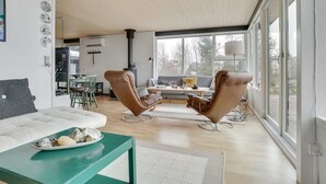 A modern living room with a large window, a sofa, two leather armchairs, a coffee table, and a dining area in the background.