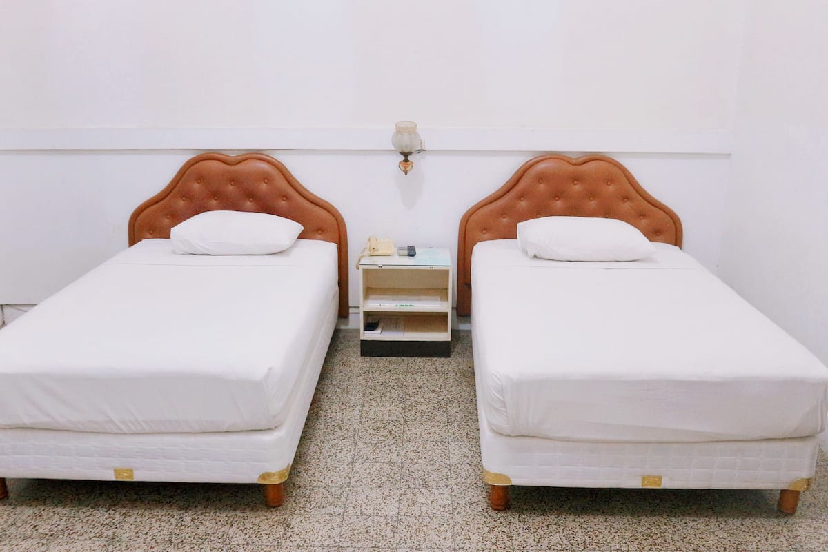 Quarto com duas camas de solteiro, uma mesinha de cabeceira entre elas e uma luminária fixada na parede.