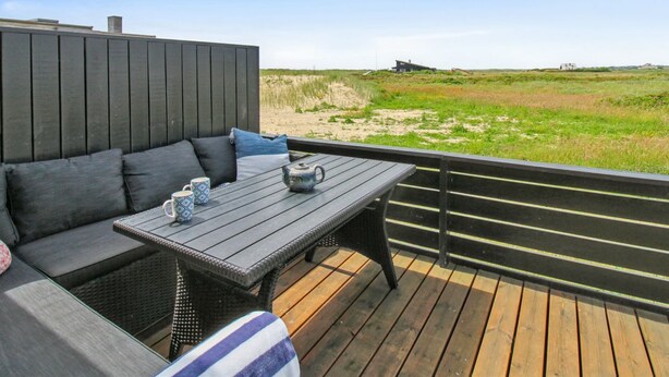 Une terrasse avec un canapé et une table, donnant sur une plage bordée de dunes et sur un bâtiment situé au loin.