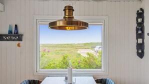 Un coin repas avec une table et des chaises, un luminaire suspendu en laiton, une fenêtre donnant sur un paysage et une étagère ornée d’objets décoratifs.