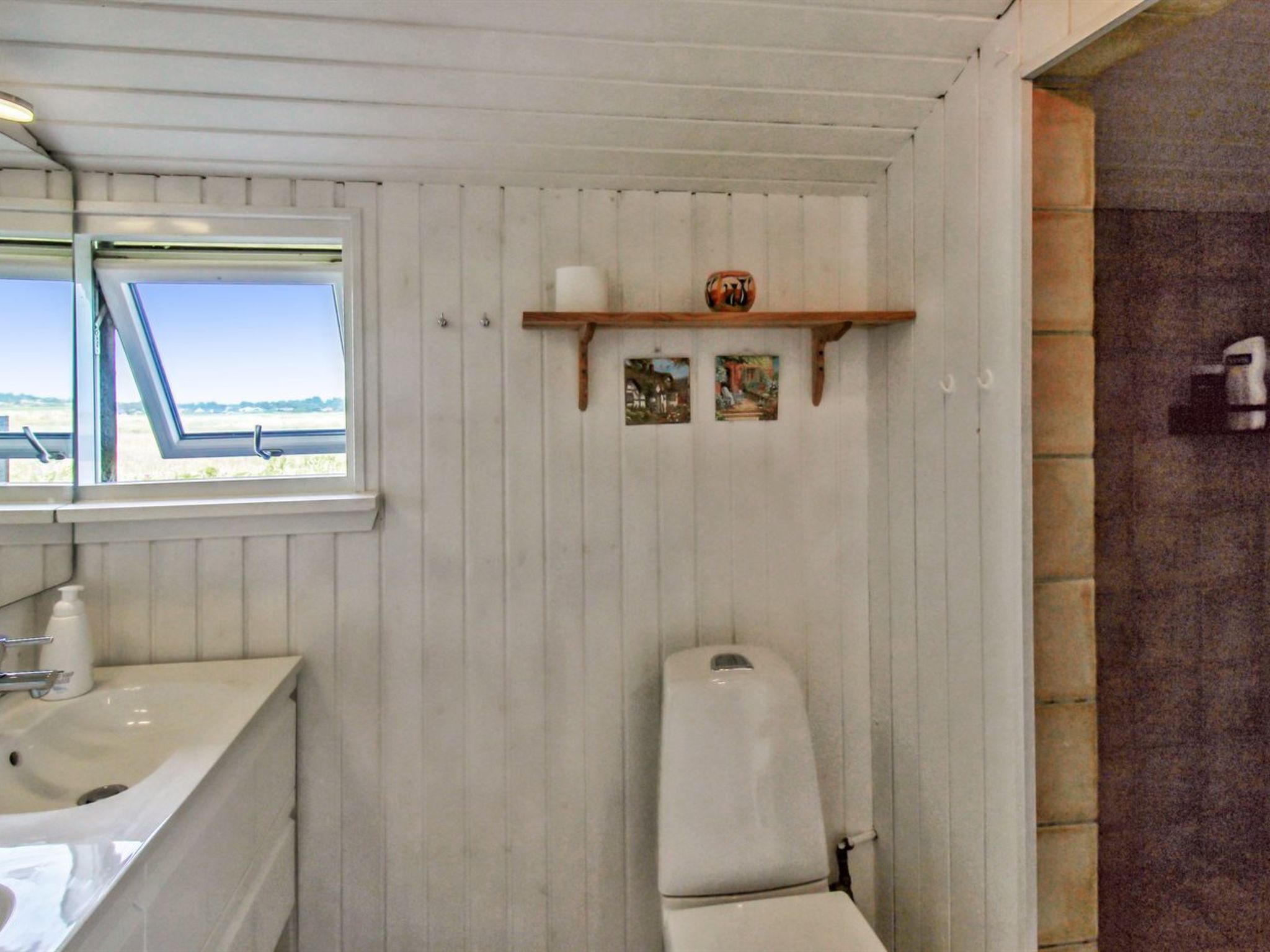 A bathroom with a window, a sink, a toilet, and a shelf with pictures.