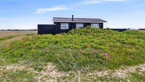 Une maison noire avec un toit en tuiles et une cheminée, entourée d’un champ de fleurs roses et d’arbustes verts.
