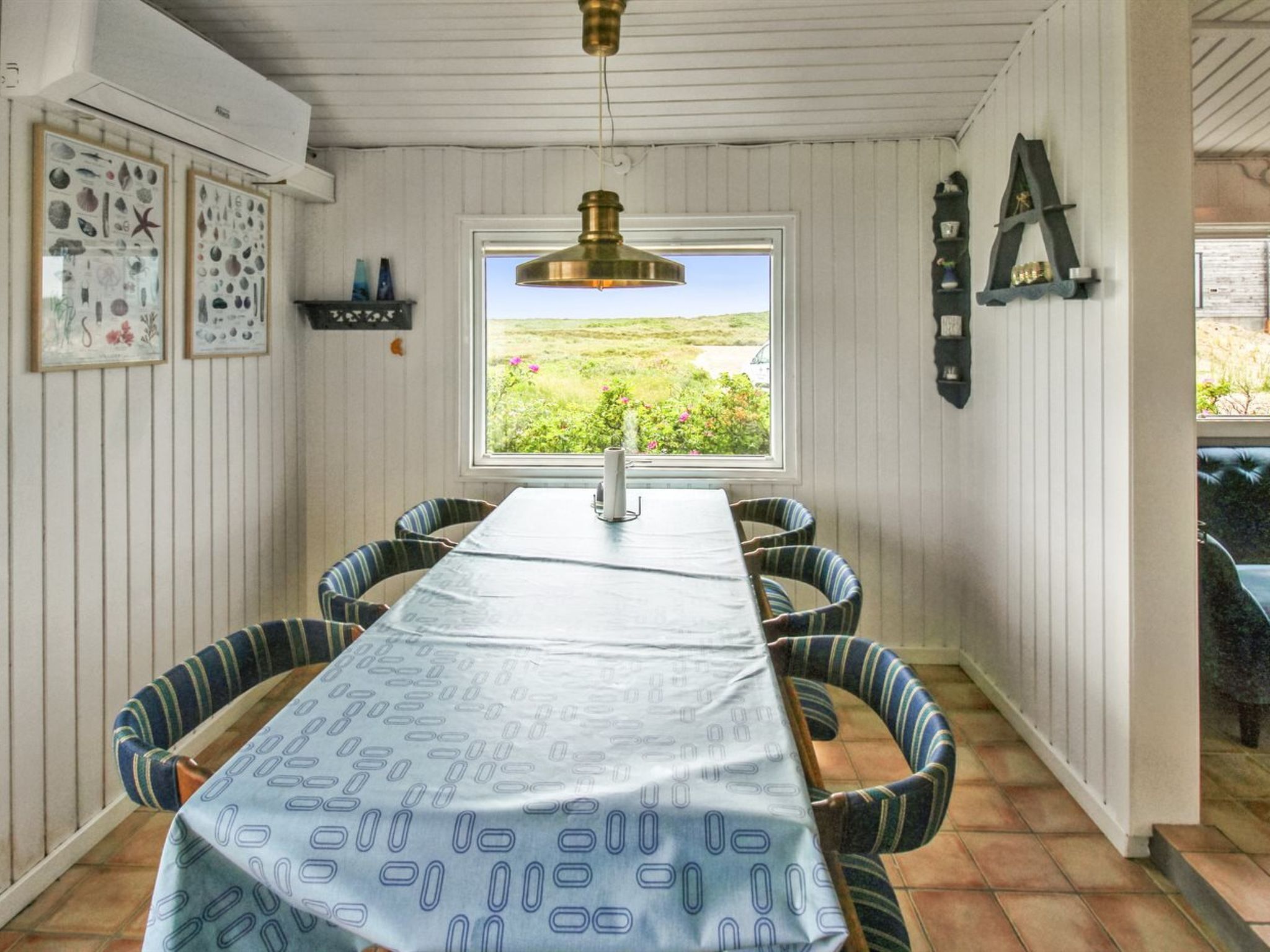 A dining area with a long table, striped chairs, a hanging light fixture, and framed artwork on the walls.