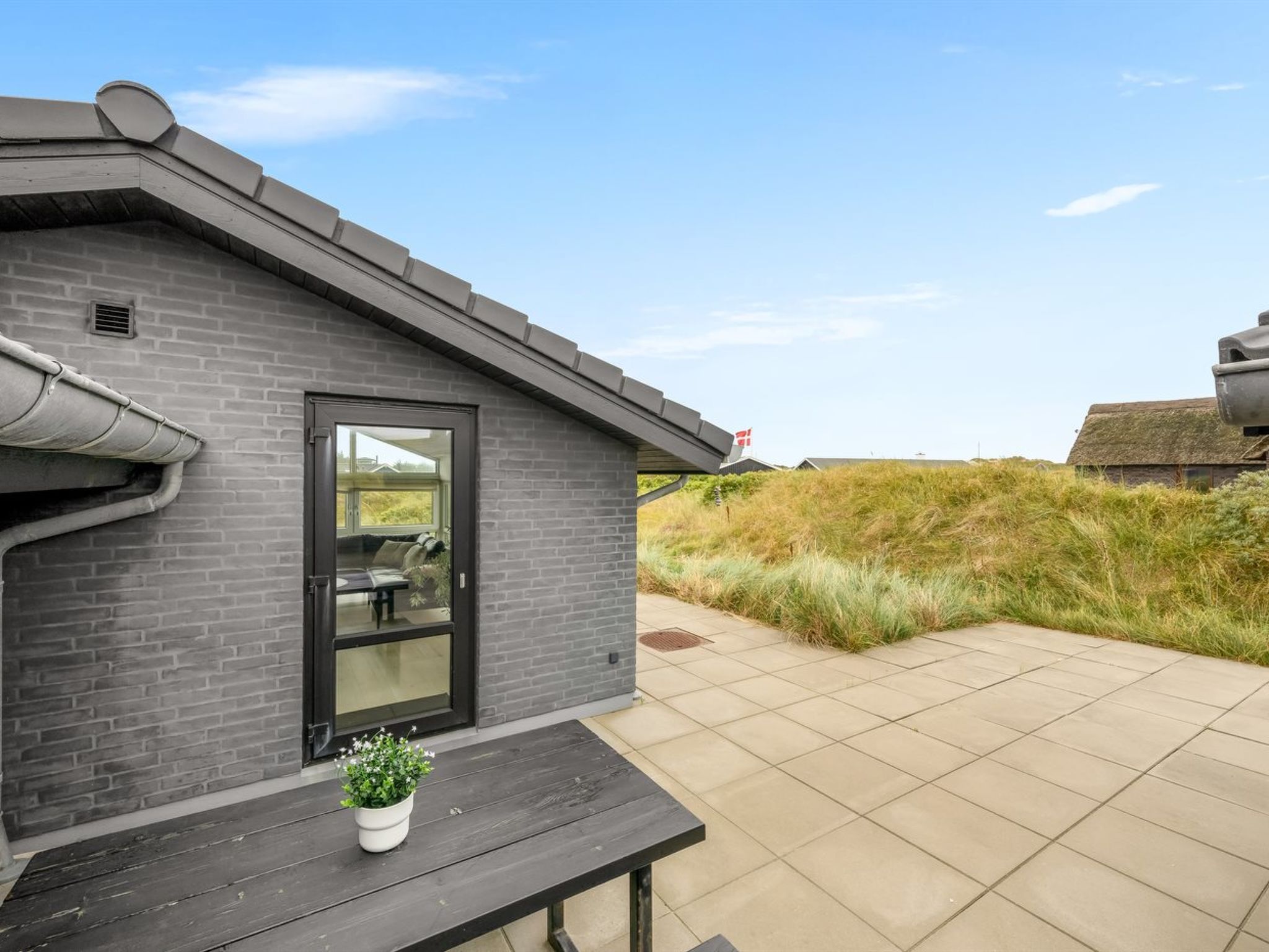 Een modern gebouw met een donkergrijze bakstenen gevel, een glazen deur en een kleine plant in een pot op een houten bankje.