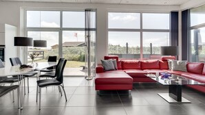 Un salon moderne avec un canapé d’angle rouge, une table basse en verre et un coin repas comprenant une table et des chaises blanches. De grandes fenêtres offrent une vue sur l’extérieur.