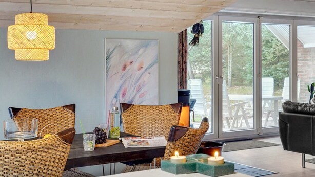 Un coin repas avec des chaises en osier, une table en bois, un luminaire suspendu et une vue sur l’extérieur grâce à de grandes fenêtres.