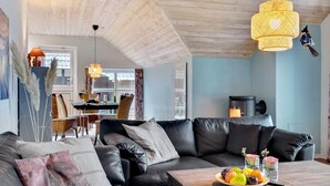 Un salon moderne avec un canapé en cuir noir, une table basse en bois sur laquelle sont disposés des fruits, et un coin repas à l'arrière-plan.