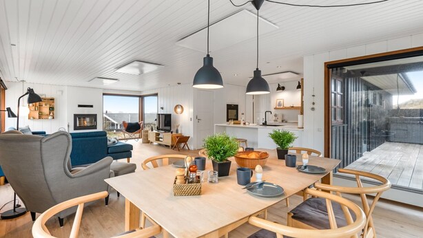 A modern living room with a wooden dining table, chairs, and pendant lights. There is a sofa, a TV, and a kitchen area visible in the background.