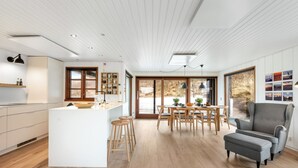A modern kitchen with a central island, wooden dining table, and bar stools. There is a large window providing a view of the outdoors.