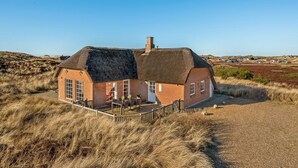 Una casa con techo de paja y un patio cercado, rodeada de pastizales secos y otras casas al fondo.