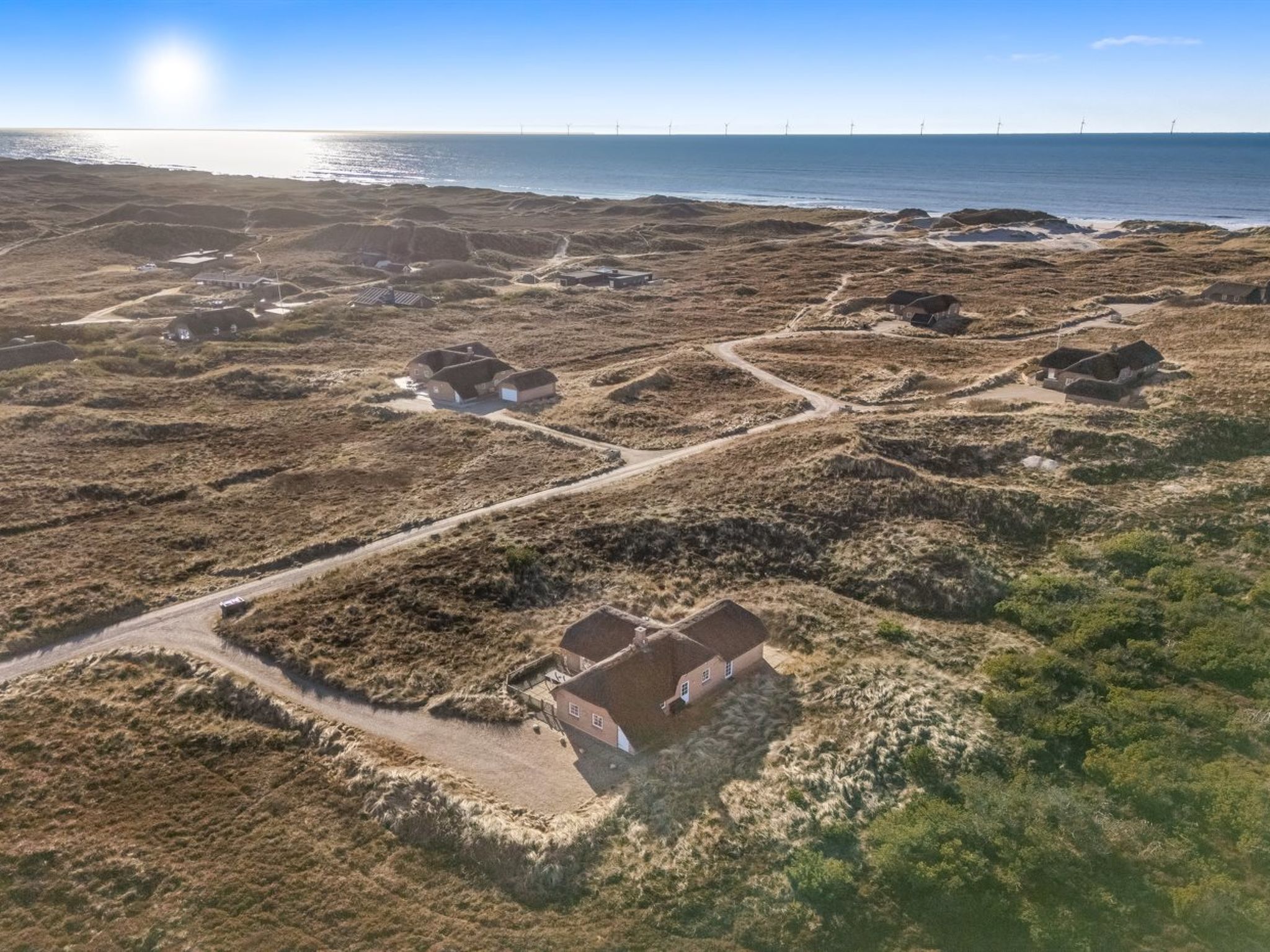 Luchtbeeld van een kustgebied met verschillende huizen, een onverharde weg en windturbines in de verte.